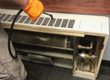 A maintenance worker in orange gloves is using a cleaning tool to service a dirty fan coil unit.