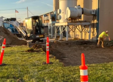 An image of two construction workers and a small excavator working outside a commercial building.